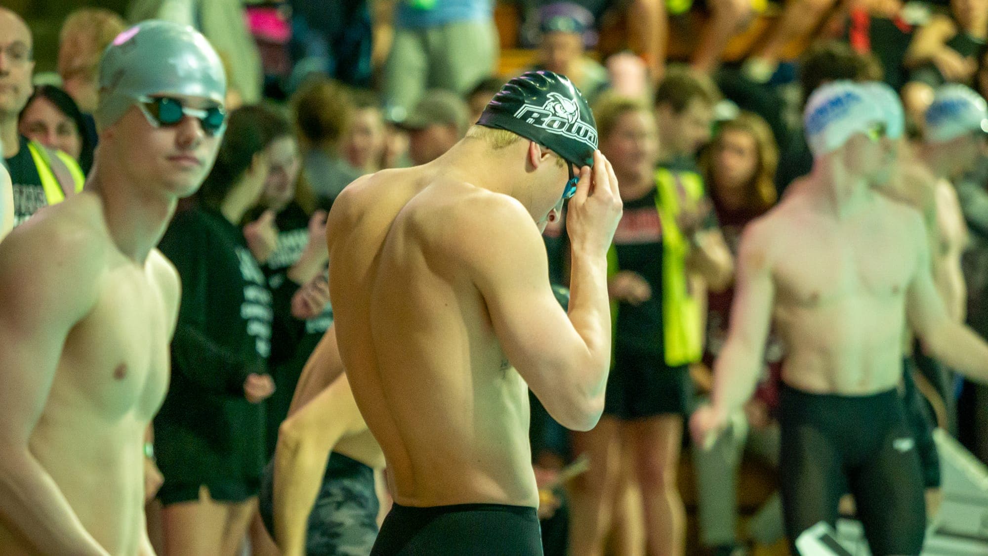 Swimmer adjusting goggles under a Rowan cap behind the blocks, teammates and crowd filling the pool deck