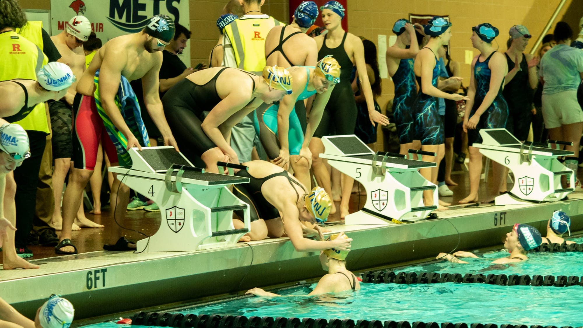 Pool deck before a relay start, swimmers climbing the numbered blocks as backstrokers wait in the water
