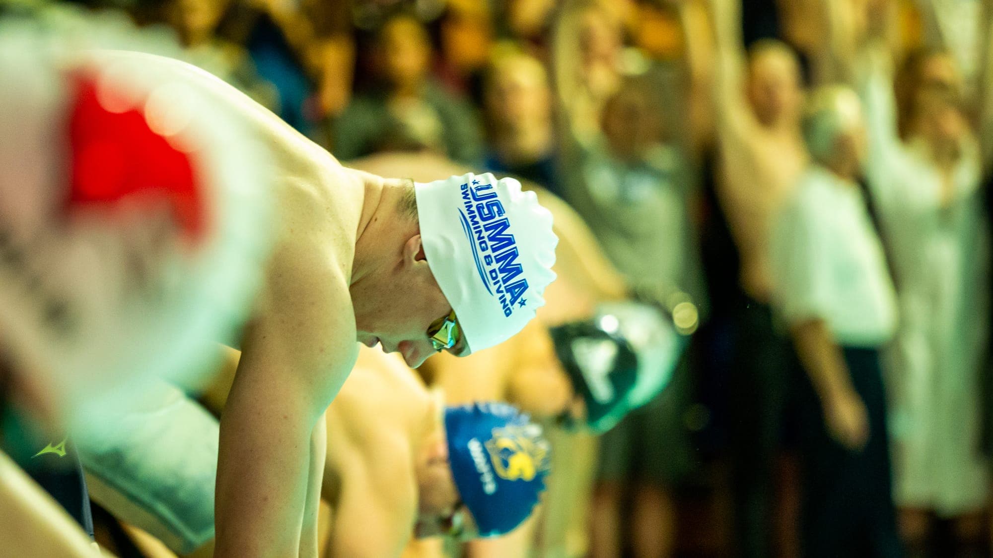 Row of swimmers bowed over the blocks before the start, a USMMA cap sharp against a blurred crowd