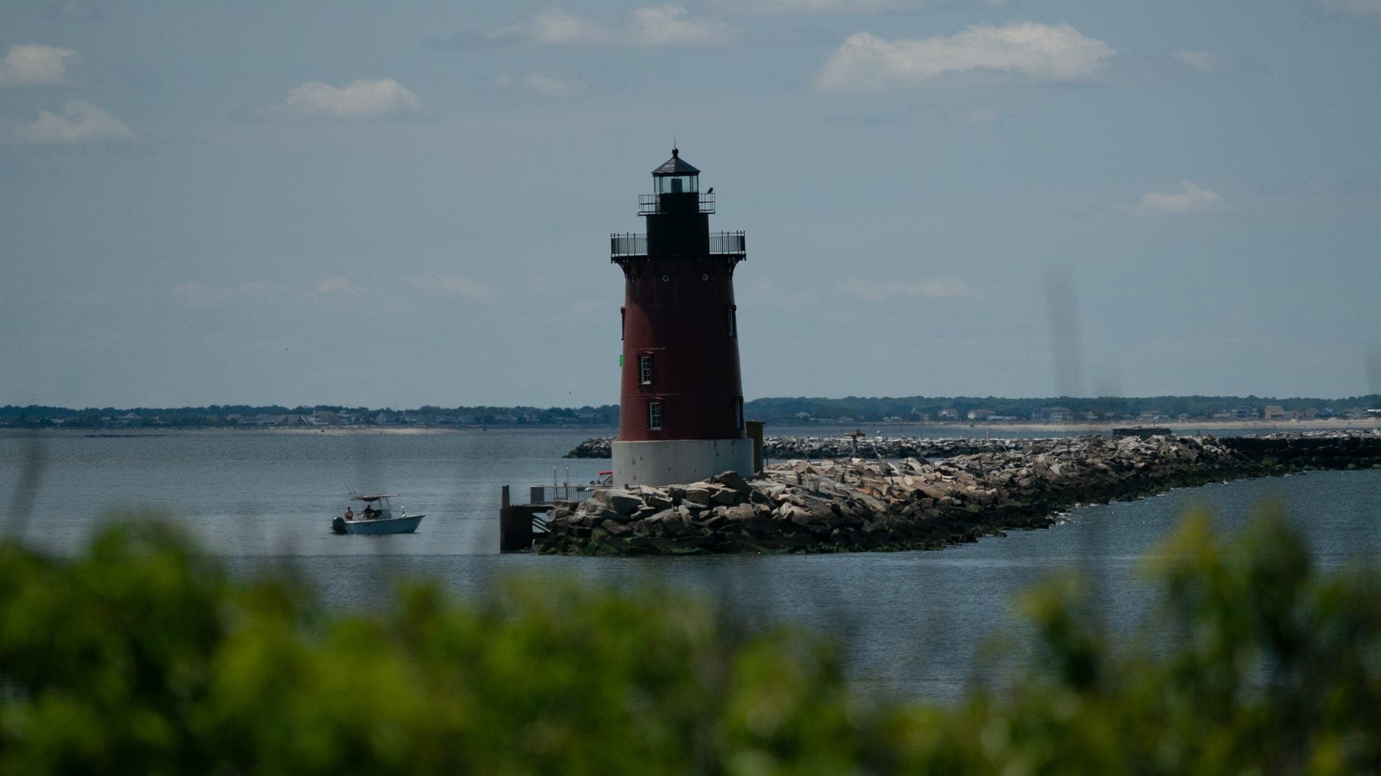 The red breakwater lighthouse stands on its stone jetty in midday haze, framed by out-of-focus green shrubs with a small fishing boat idling on the calm bay.