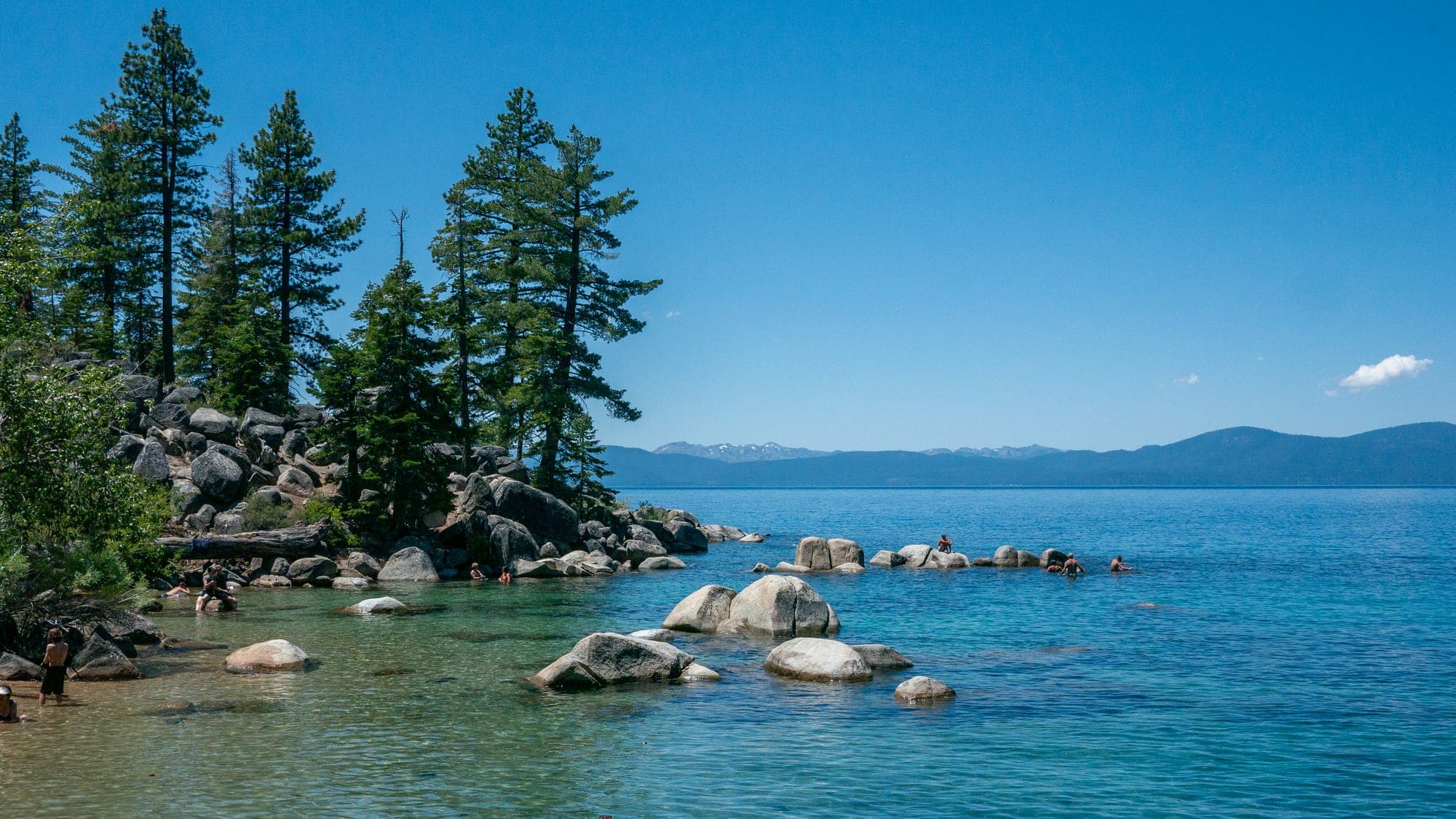 Granite boulders and pines edge a clear turquoise cove where swimmers wade among smooth rocks, with hazy mountains across the lake under bright summer light.
