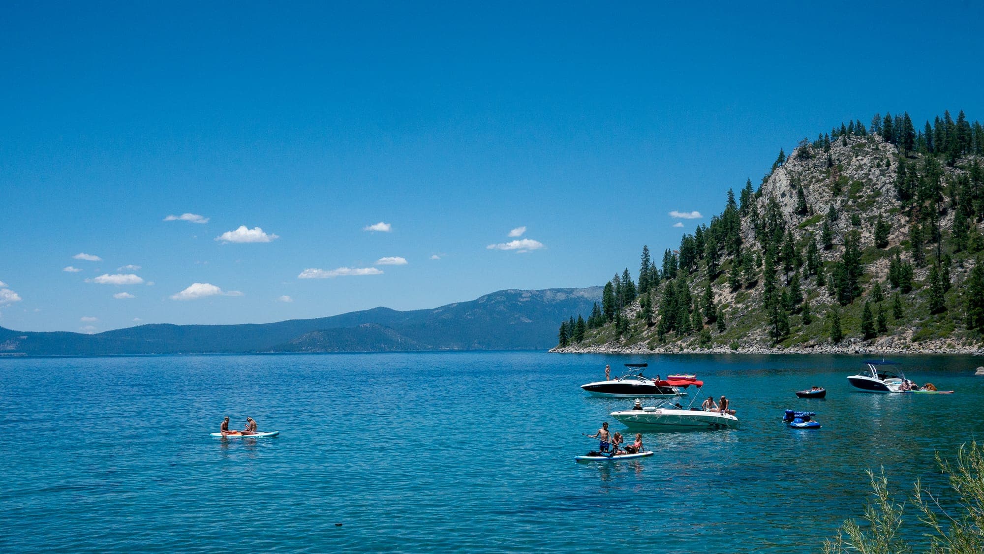 Paddleboarders and small boats drift on vivid blue alpine water beneath a rocky pine-covered headland and scattered fair-weather clouds.