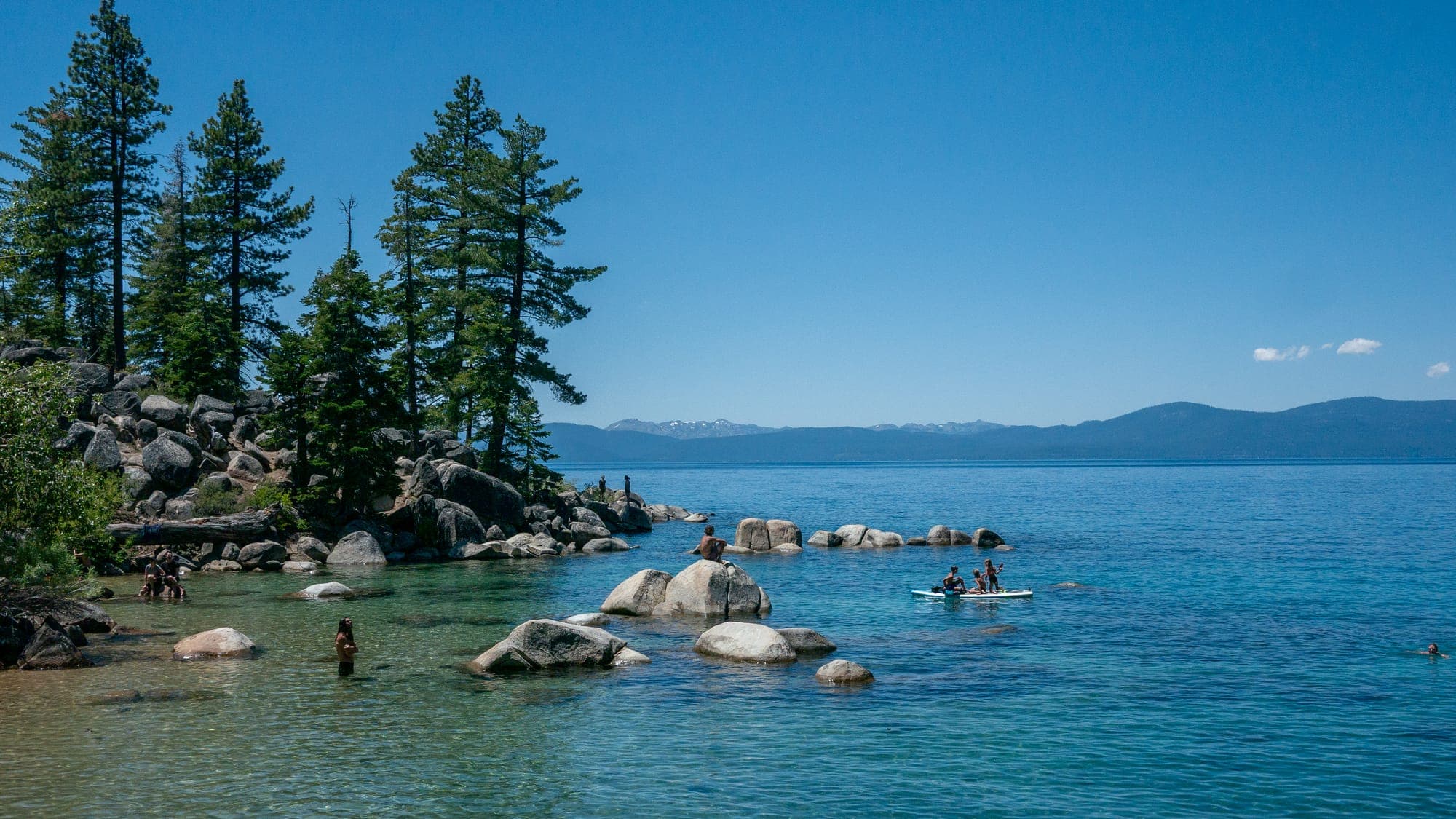 Swimmers and a loaded paddleboard scatter across a shallow boulder-strewn cove, clear green water grading to deep blue toward distant snow-flecked peaks.