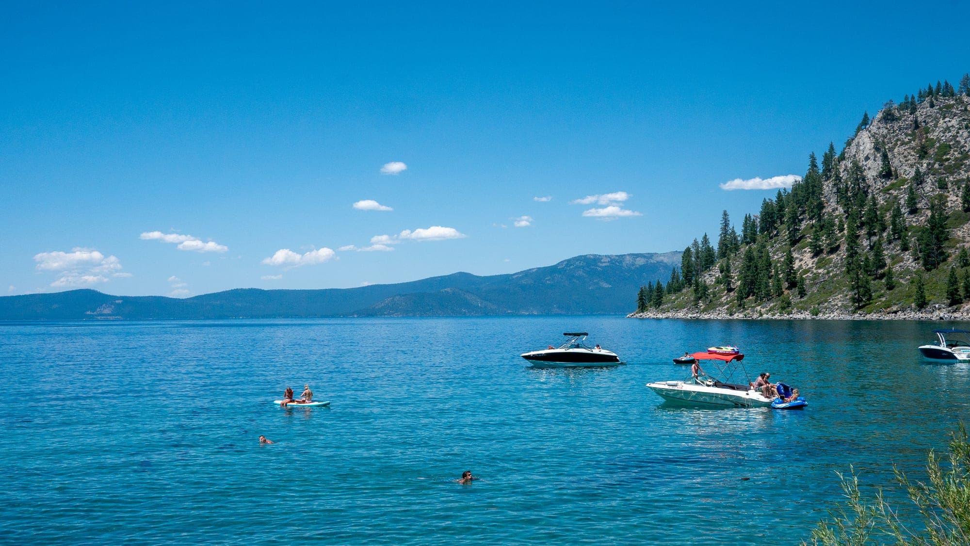 Motorboats, a paddleboarder, and swimmers idle in calm turquoise water below a steep granite slope of pines on a bright summer afternoon.
