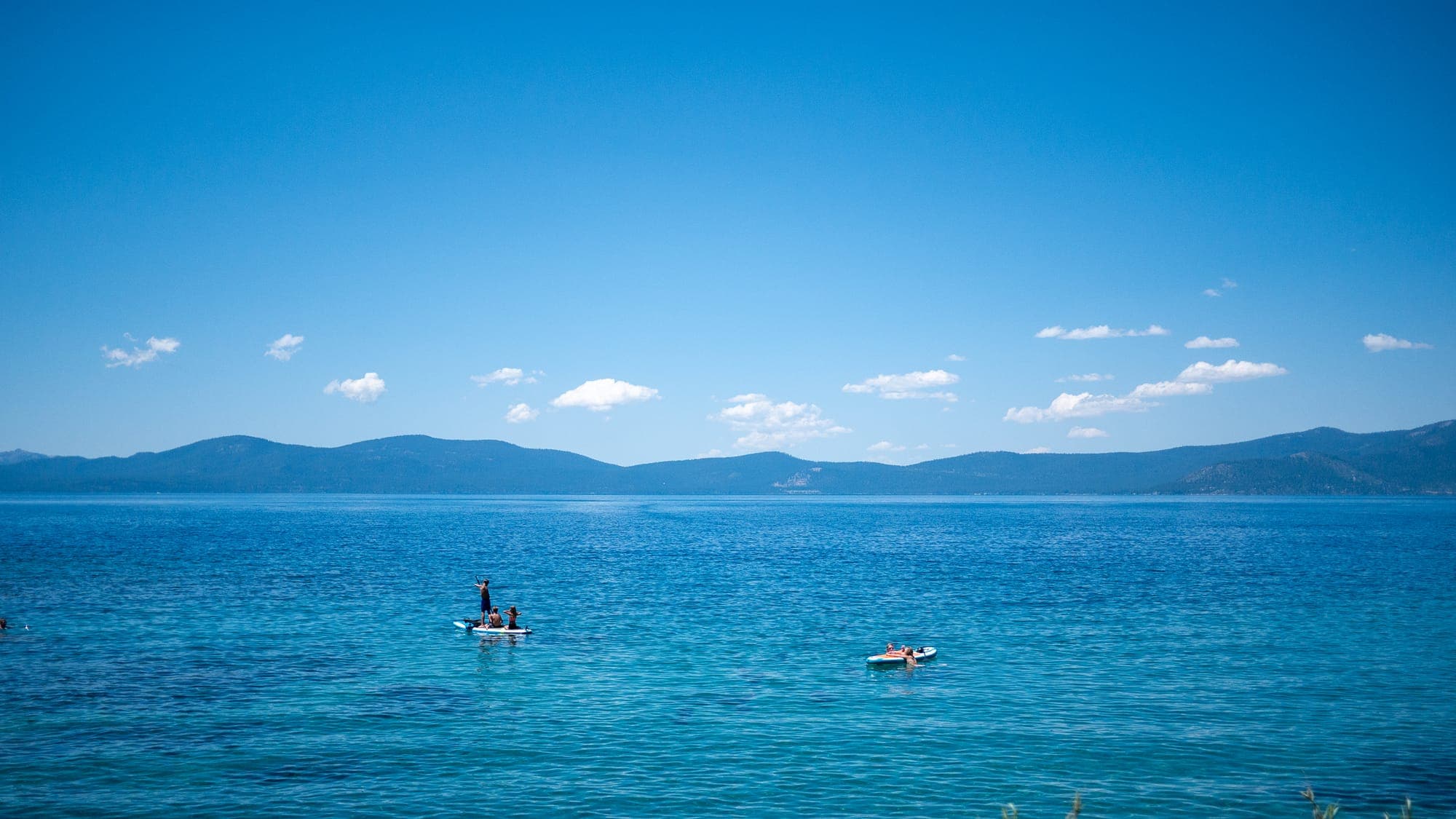 A vast plane of graded blue water and sky dwarfs two tiny paddleboarders, with a low ridge of mountains pinched along the horizon.