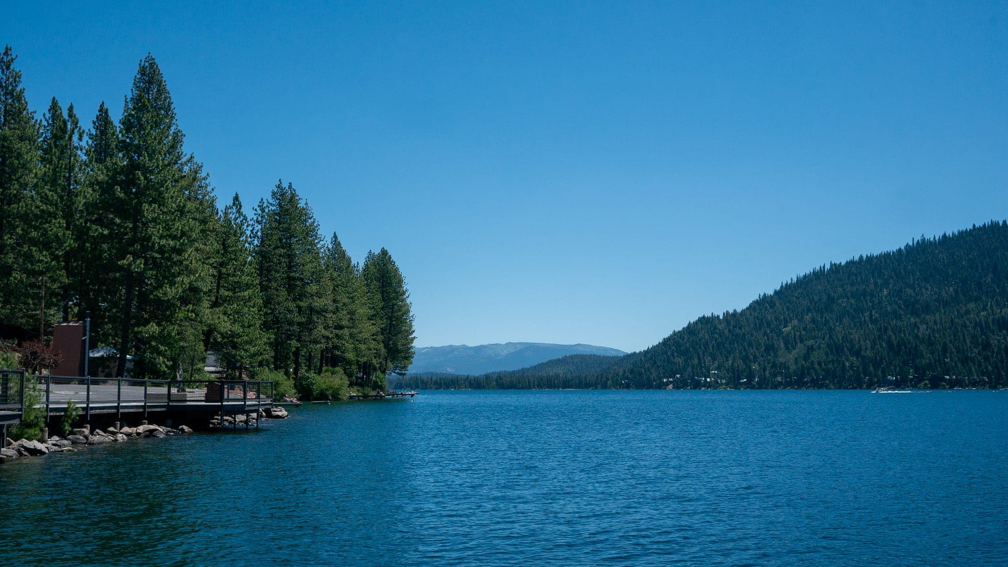 A wooden lakeside boardwalk curves beneath tall pines along still, deep blue water, a forested ridge sloping to the far shore in midday haze.