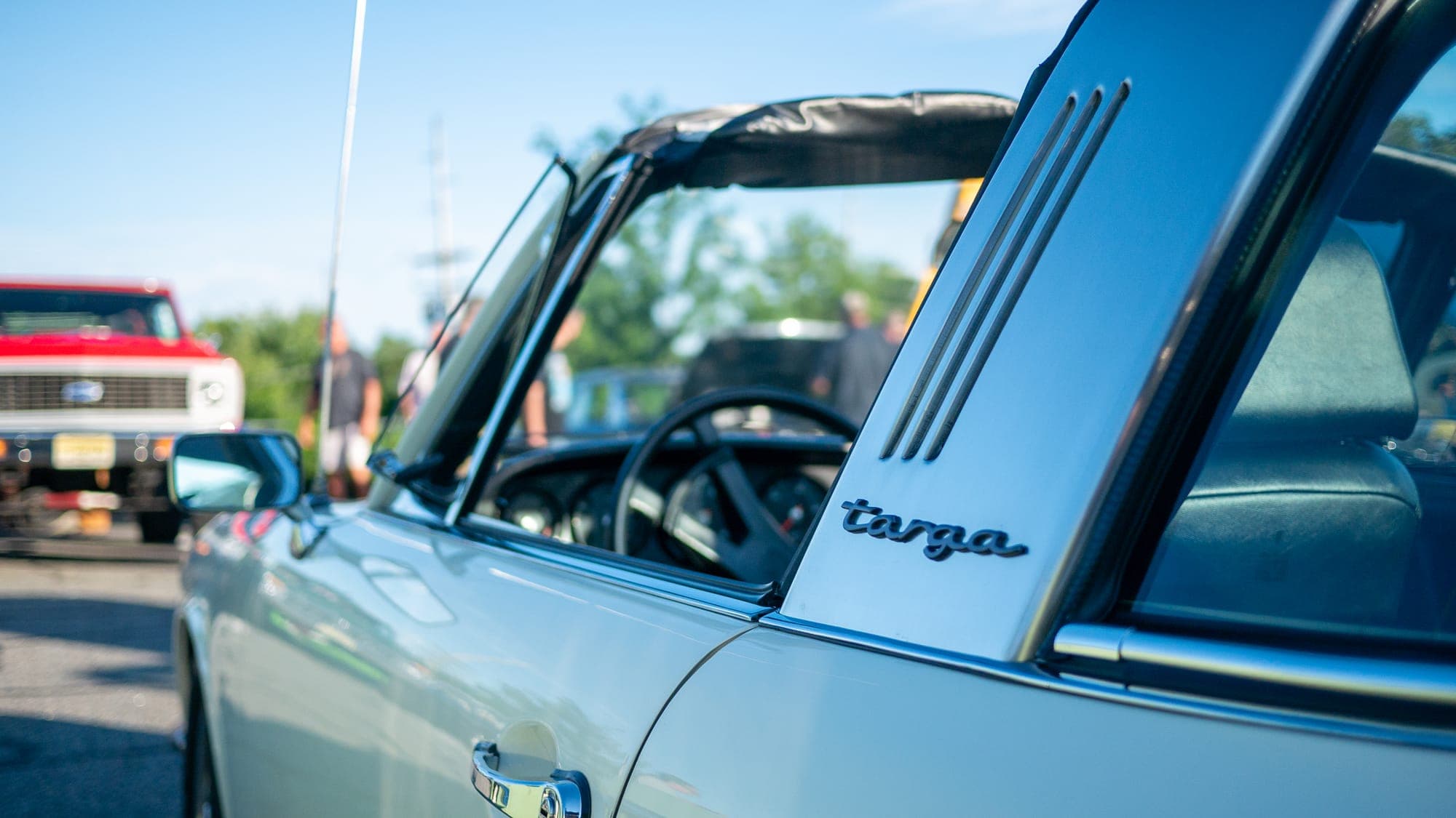 Targa script and brushed roll-bar pillar of a pale blue Porsche 911, a red pickup soft in the background.
