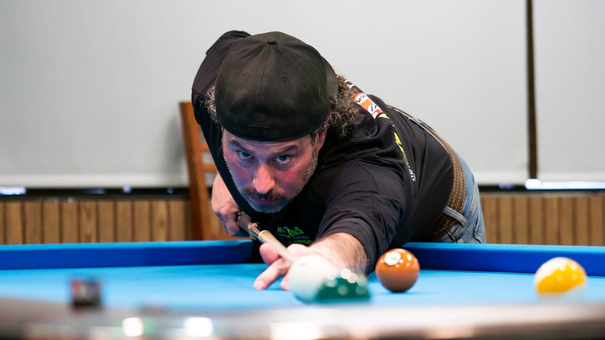 Pool player in a backwards cap stretching across the table mid-stroke, cue ball and orange ball sharp in the foreground