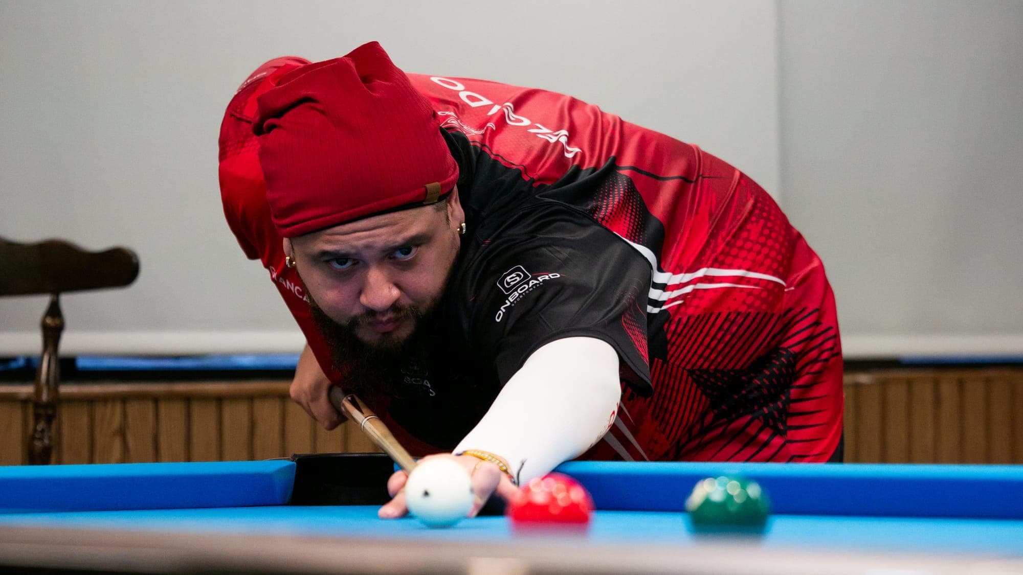 Pool player in a red headwrap and jersey bridging a shot, white cue ball balanced at his fingertips