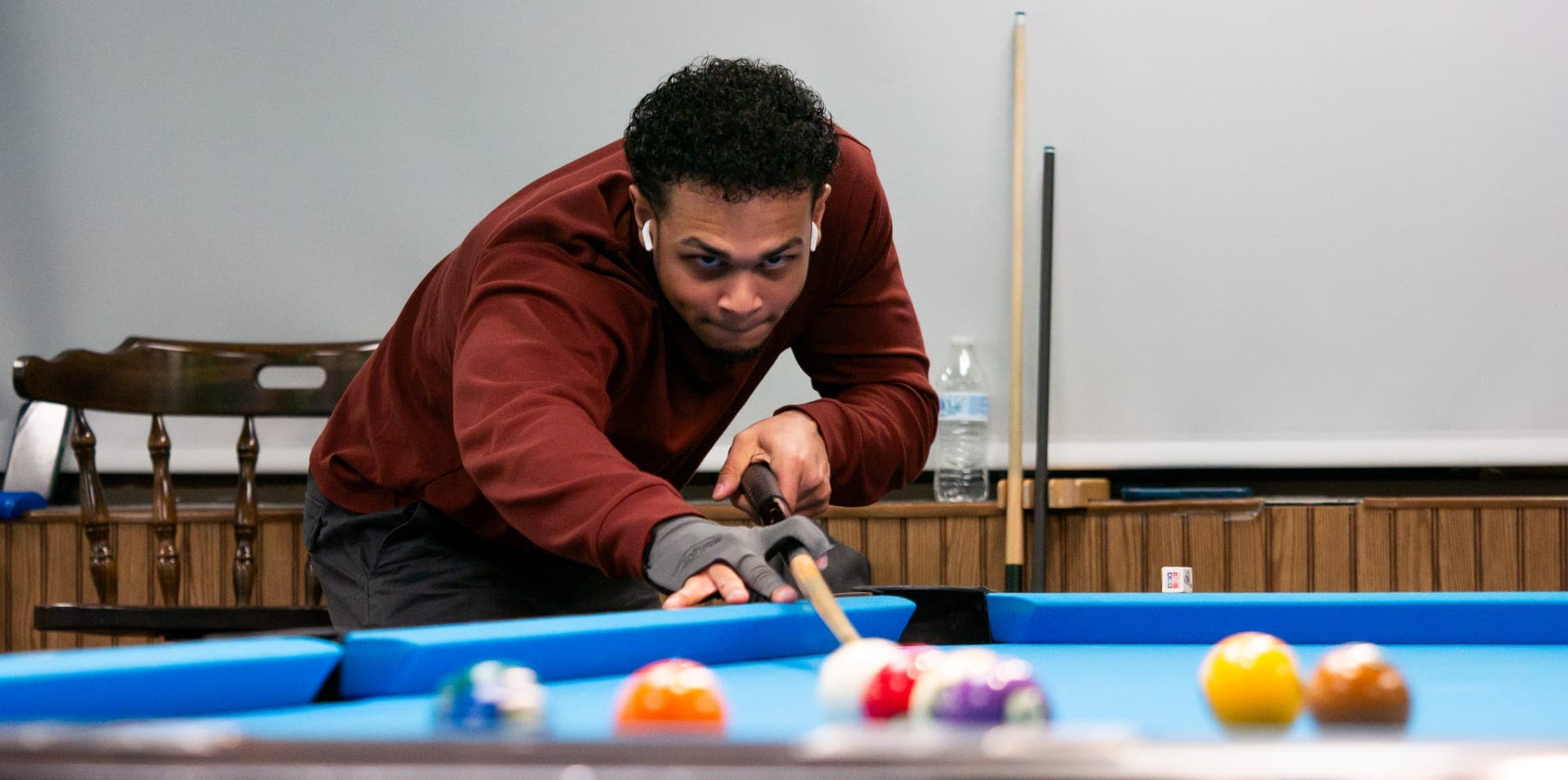 Pool player in maroon lining up a shot behind a scatter of colored balls on bright blue felt