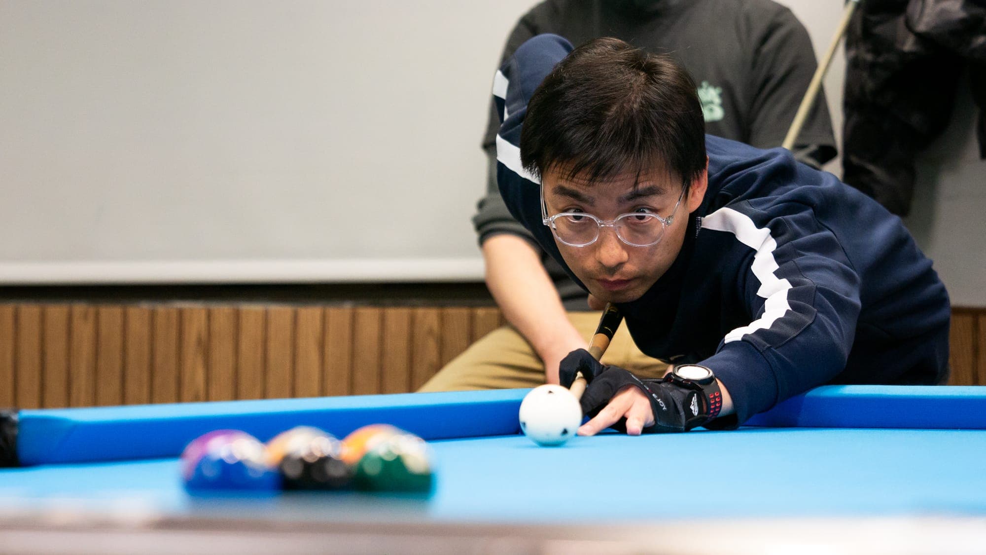 Billiards player in glasses and a navy track jacket cradling the cue over the white ball, racked balls blurred nearby