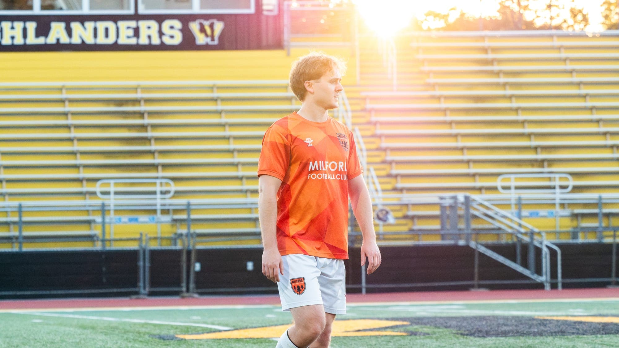 Soccer player in an orange Milford Football Club kit walking the pitch at sunset, empty yellow bleachers glowing behind him