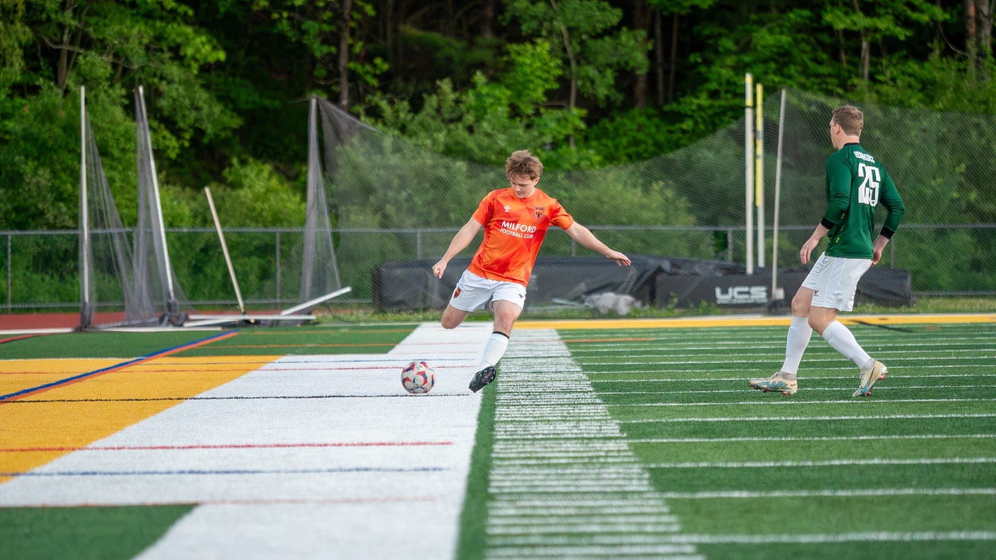 Soccer player in orange winding up to strike the ball at the sideline as a green-shirted defender closes in