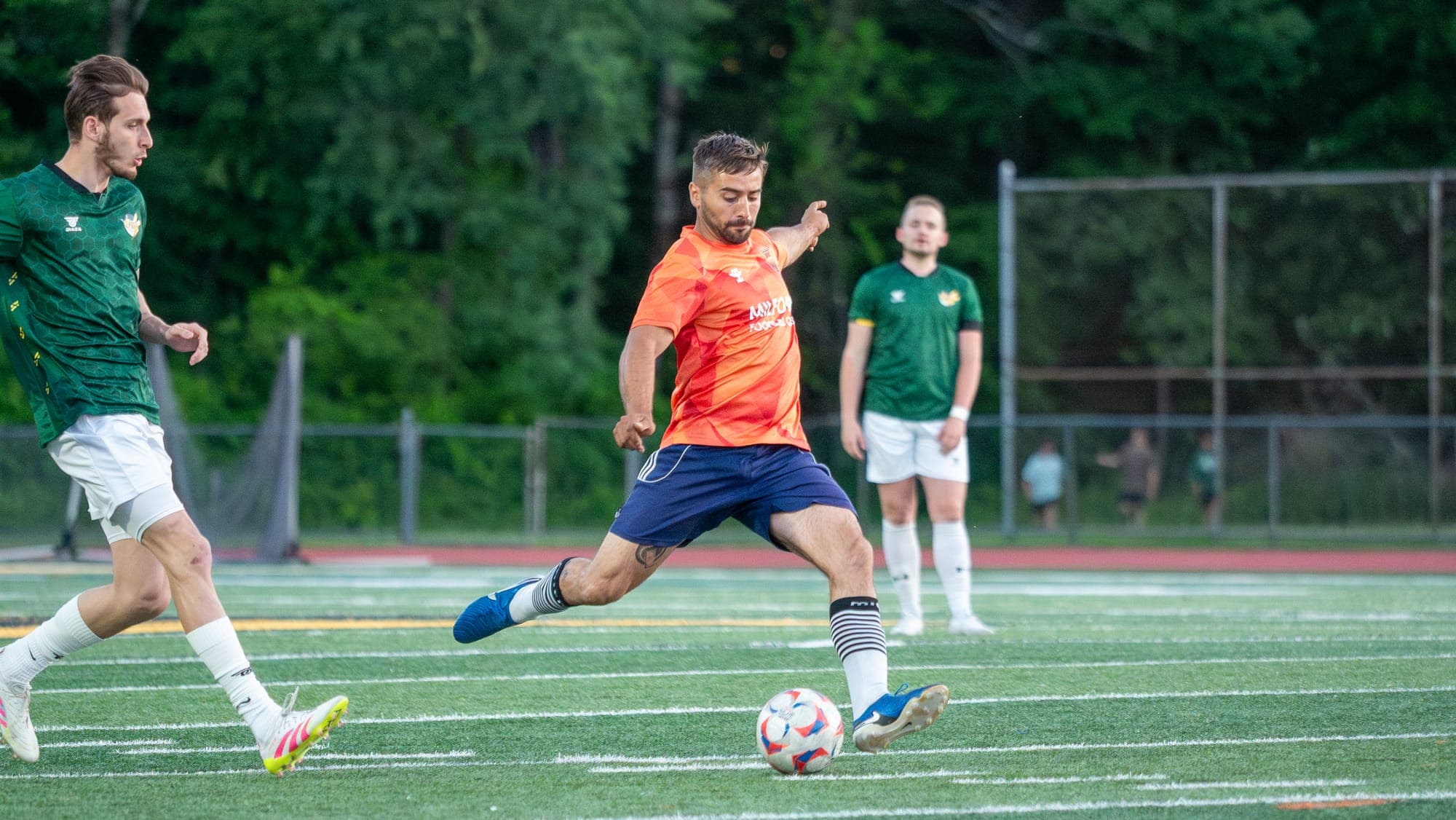 Soccer player in orange mid-kick, leg coiled over the ball as opponents in green watch the play