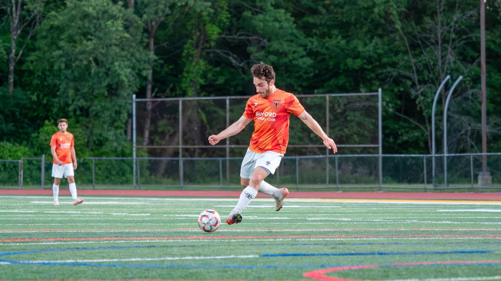 Soccer player in an orange Milford FC kit winding up to strike the ball on a green turf field