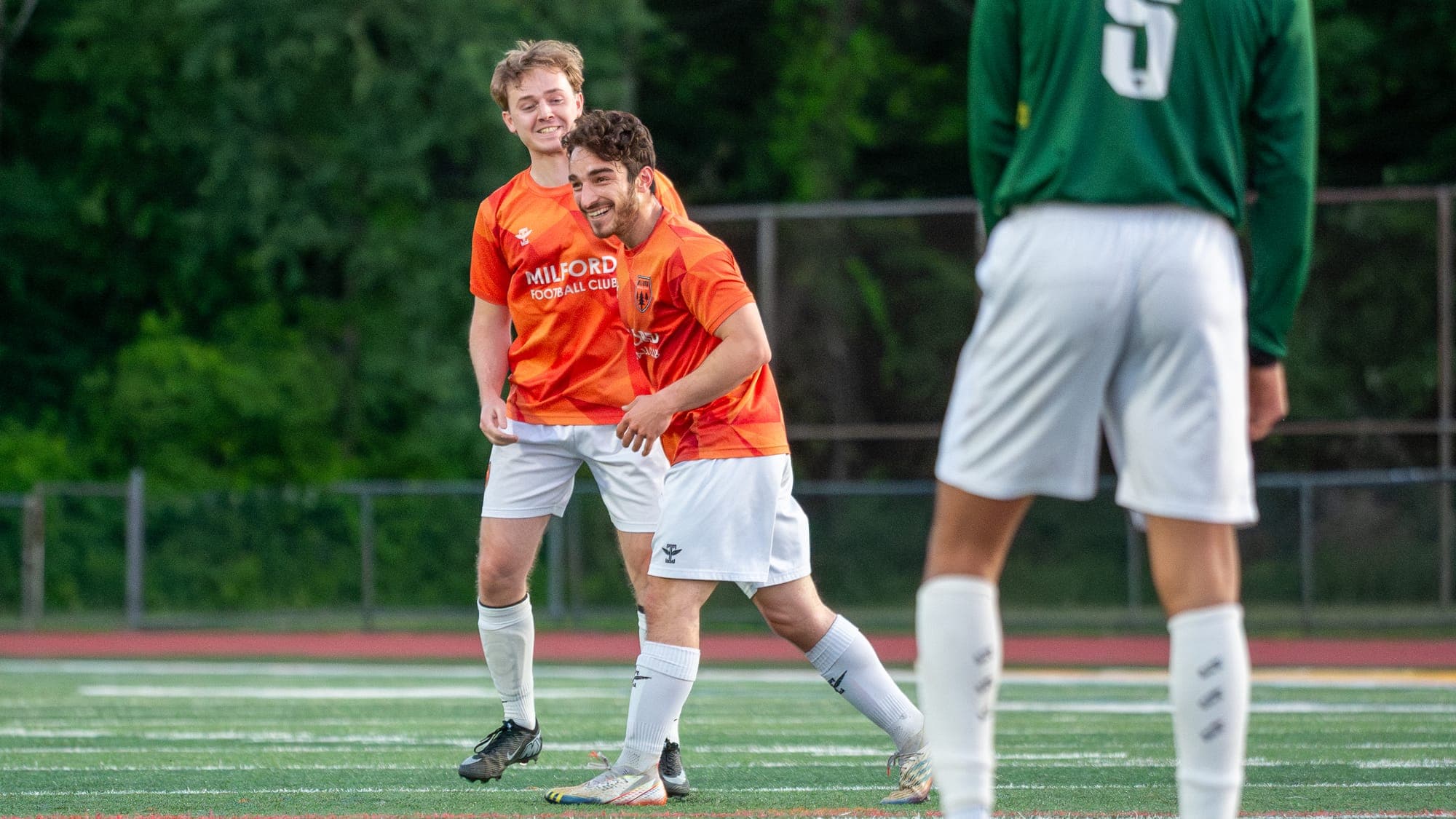 Two Milford FC teammates grinning together after a goal, framed past a defender's back