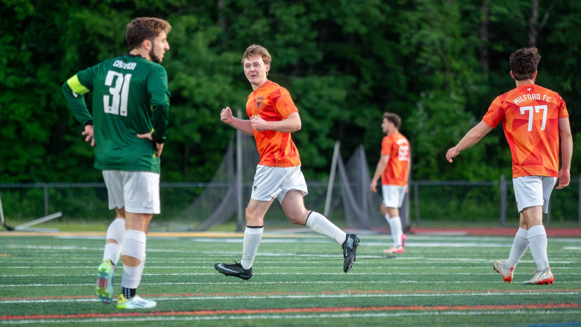 Soccer player jogging back smiling after a goal while the opposing captain stands with hands on hips