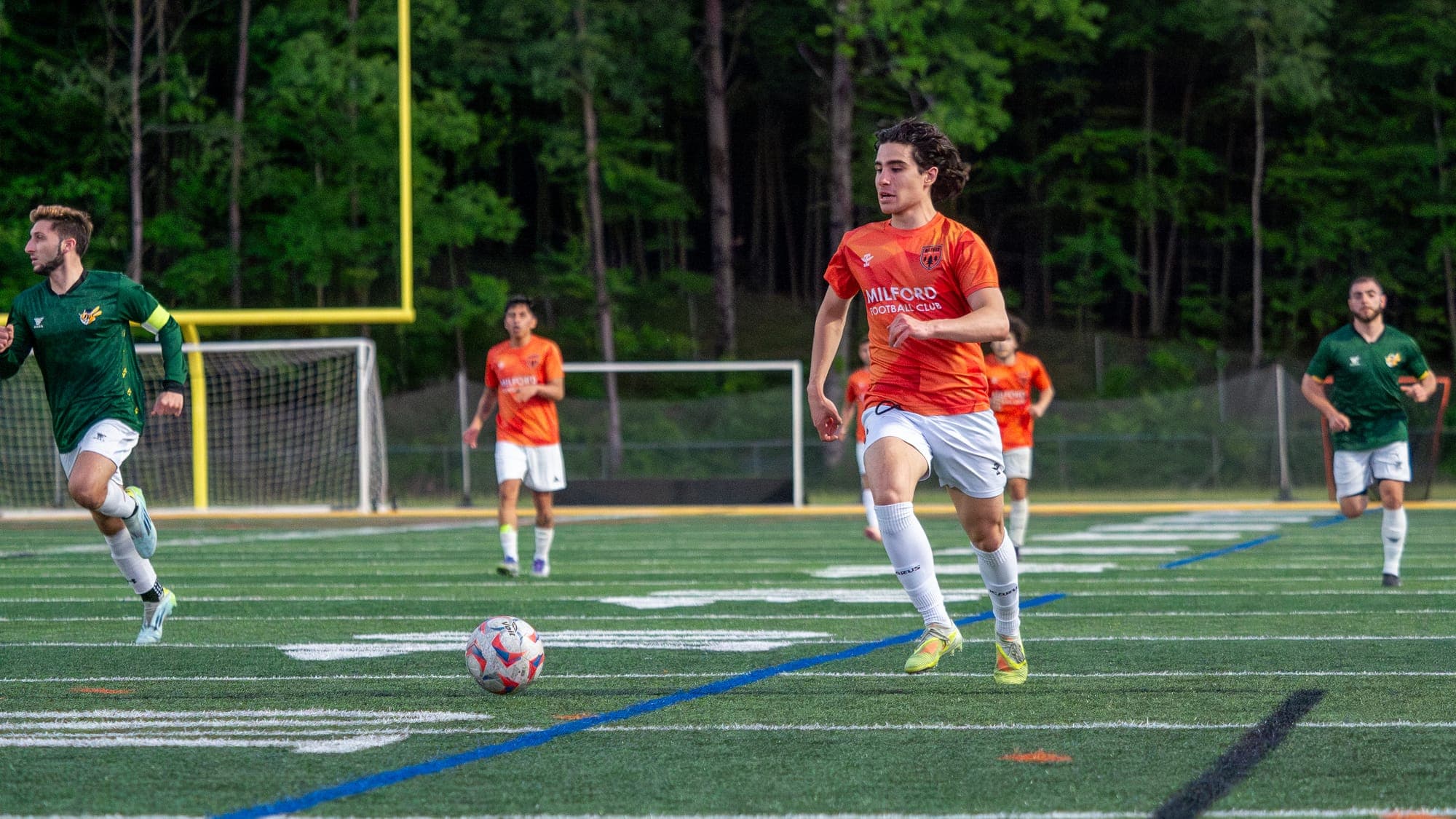 Long-haired soccer player in orange driving upfield toward a loose ball ahead of chasing opponents