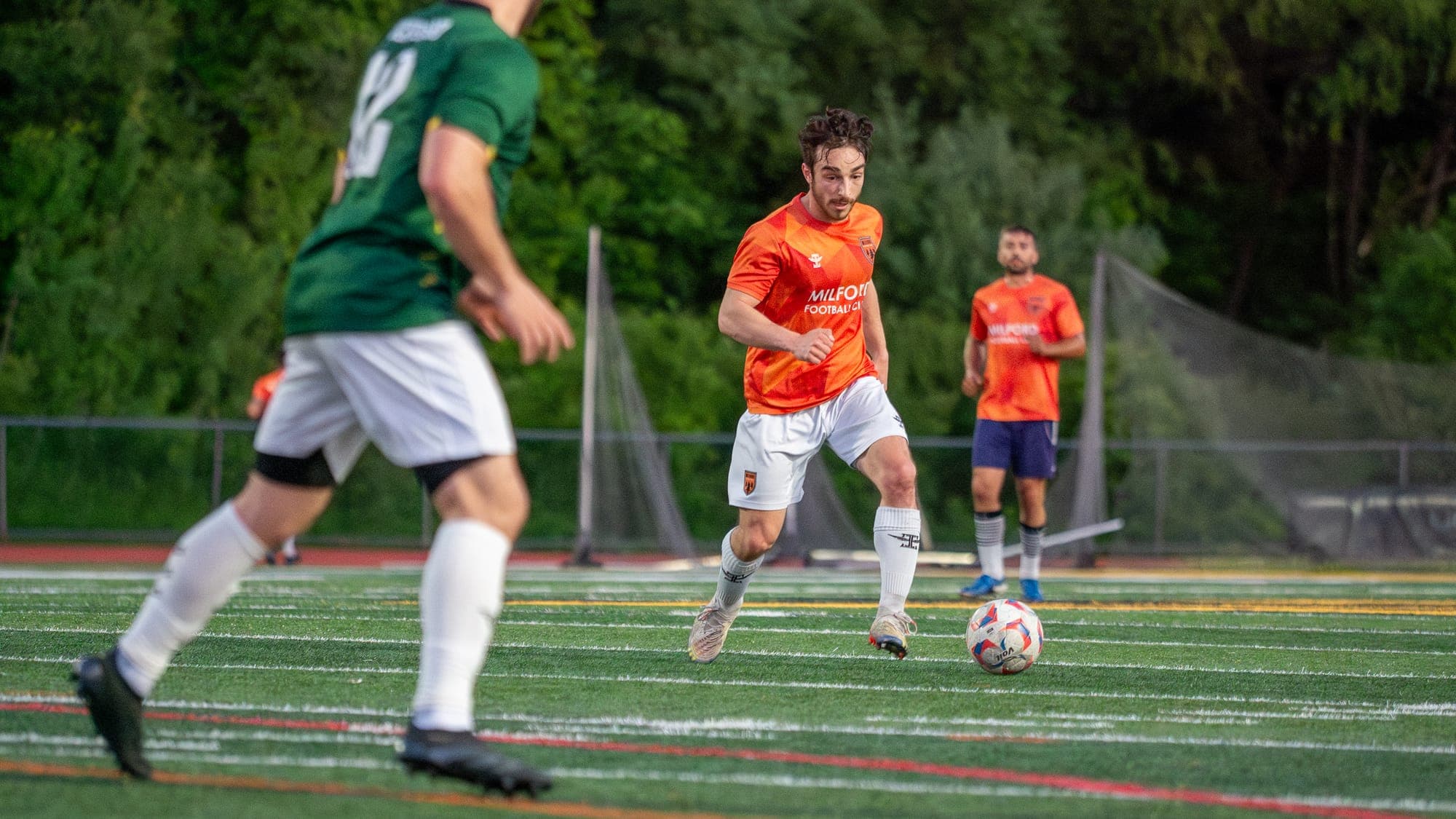 Soccer player in orange carrying the ball forward, framed through the legs of an approaching defender