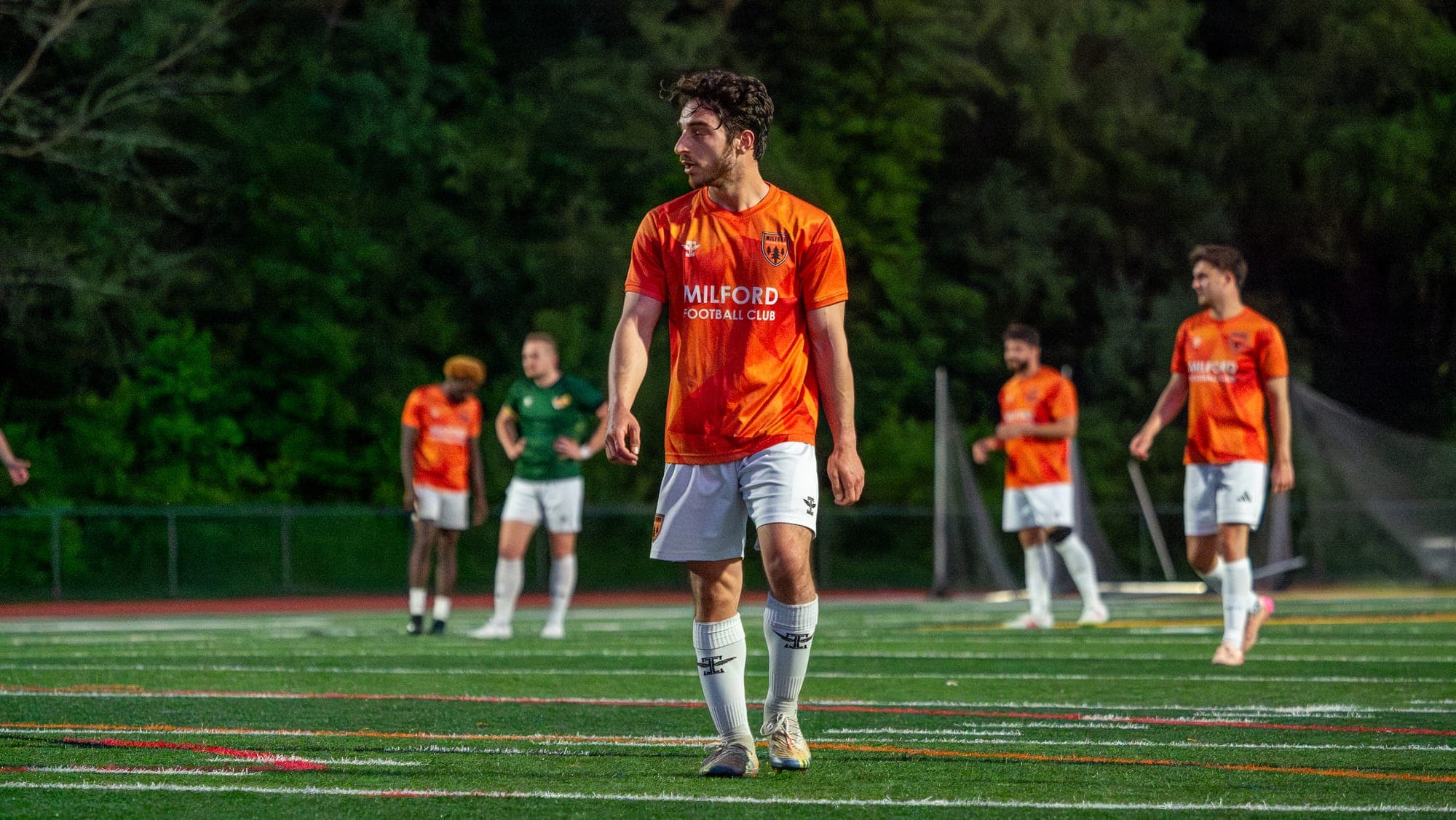 Soccer player walking the turf and glancing over his shoulder, teammates scattered behind him