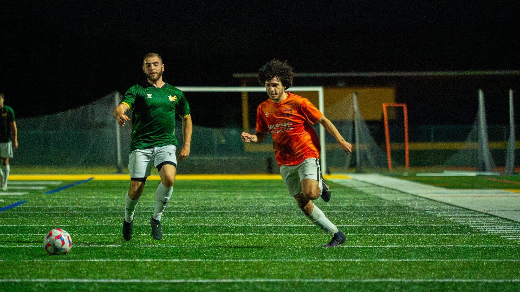 Soccer player in orange and a defender in green sprinting flat-out for the ball under night lights
