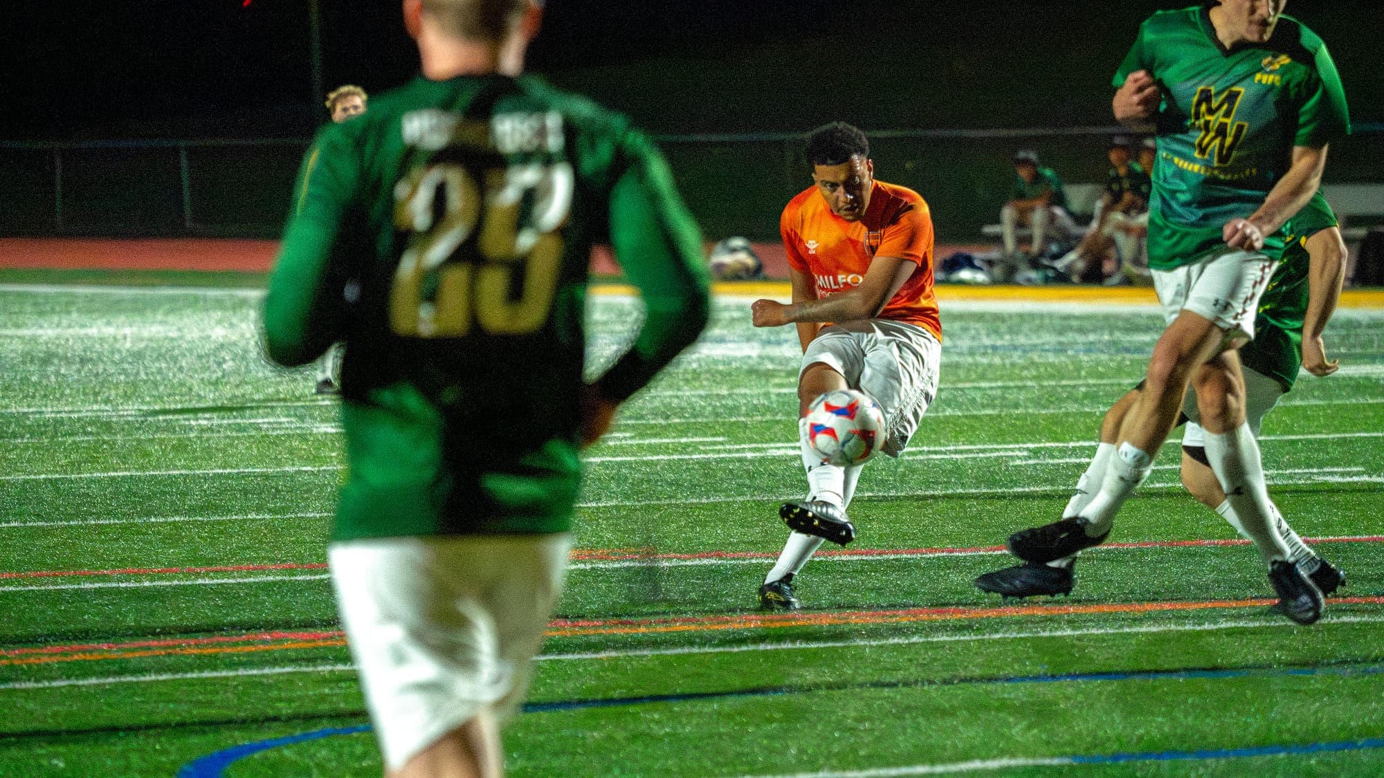 Soccer player in orange striking a shot through a crowd of defenders, framed past number 23's back