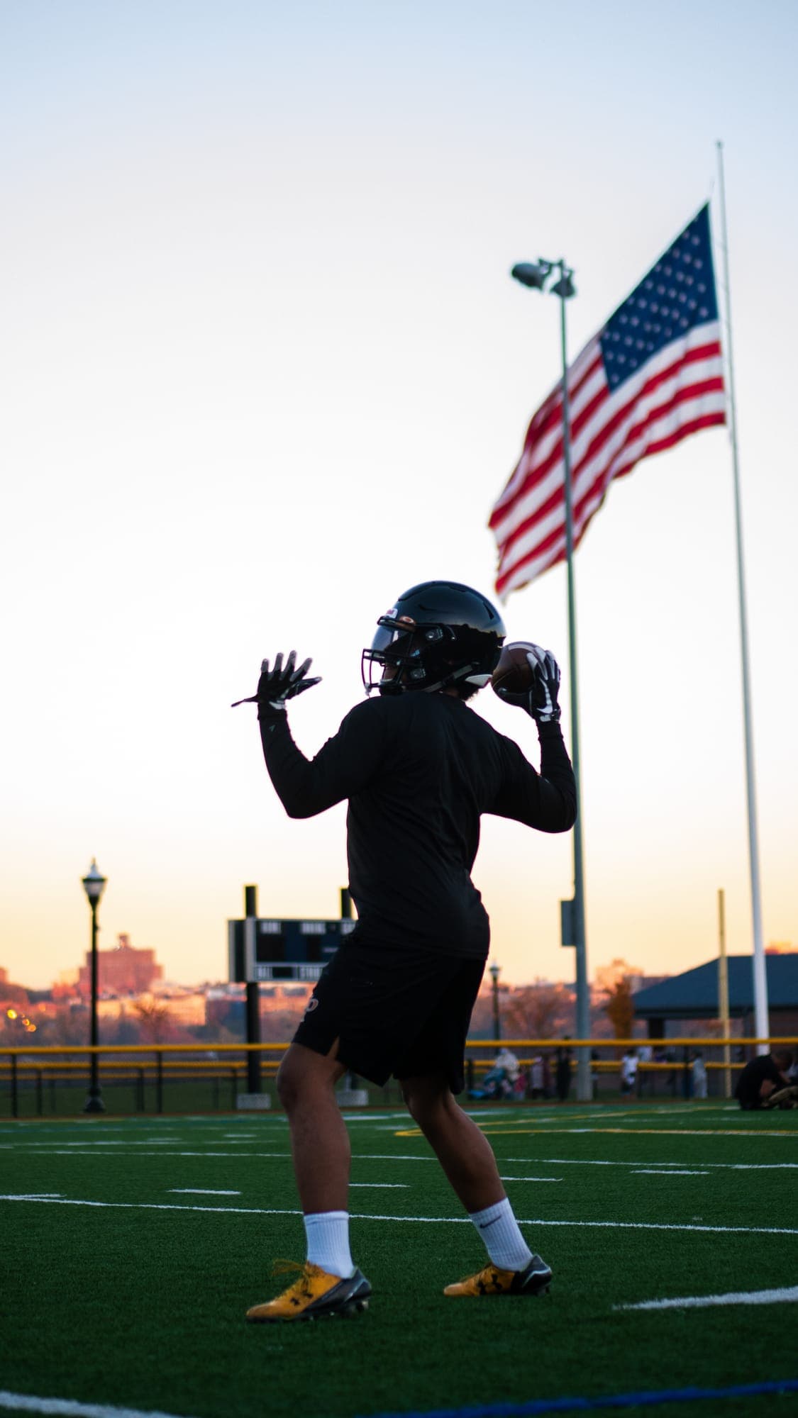 Football quarterback silhouetted at dusk, cocking the ball to throw beneath an American flag