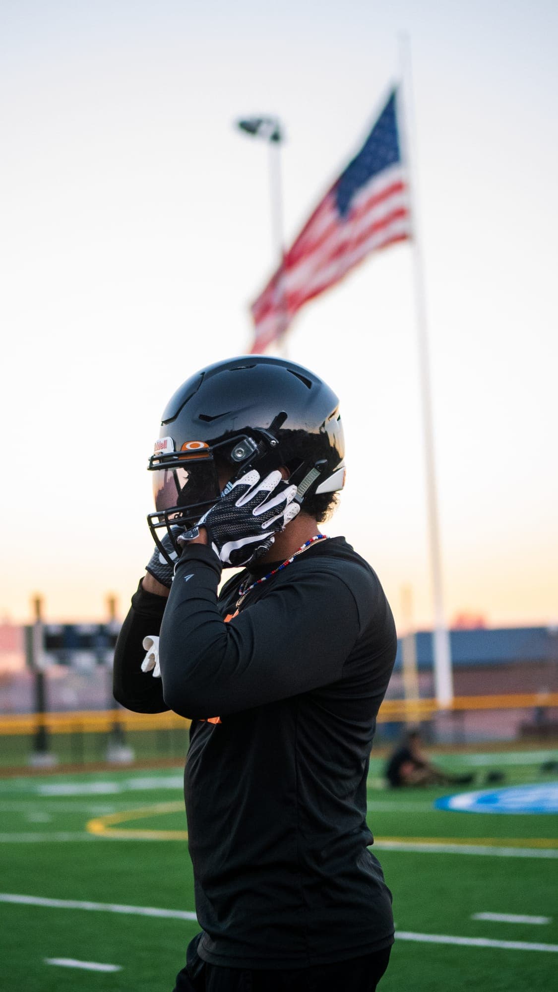 Football player fastening his helmet chinstrap at sunset, flag rippling out of focus behind him