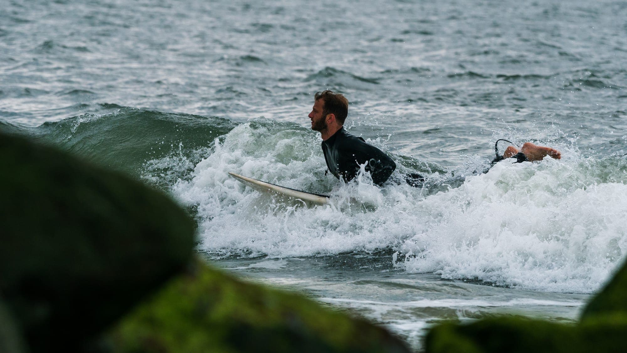 Surfer pushing through collapsing whitewater, board nose surfacing beside the mossy rocks