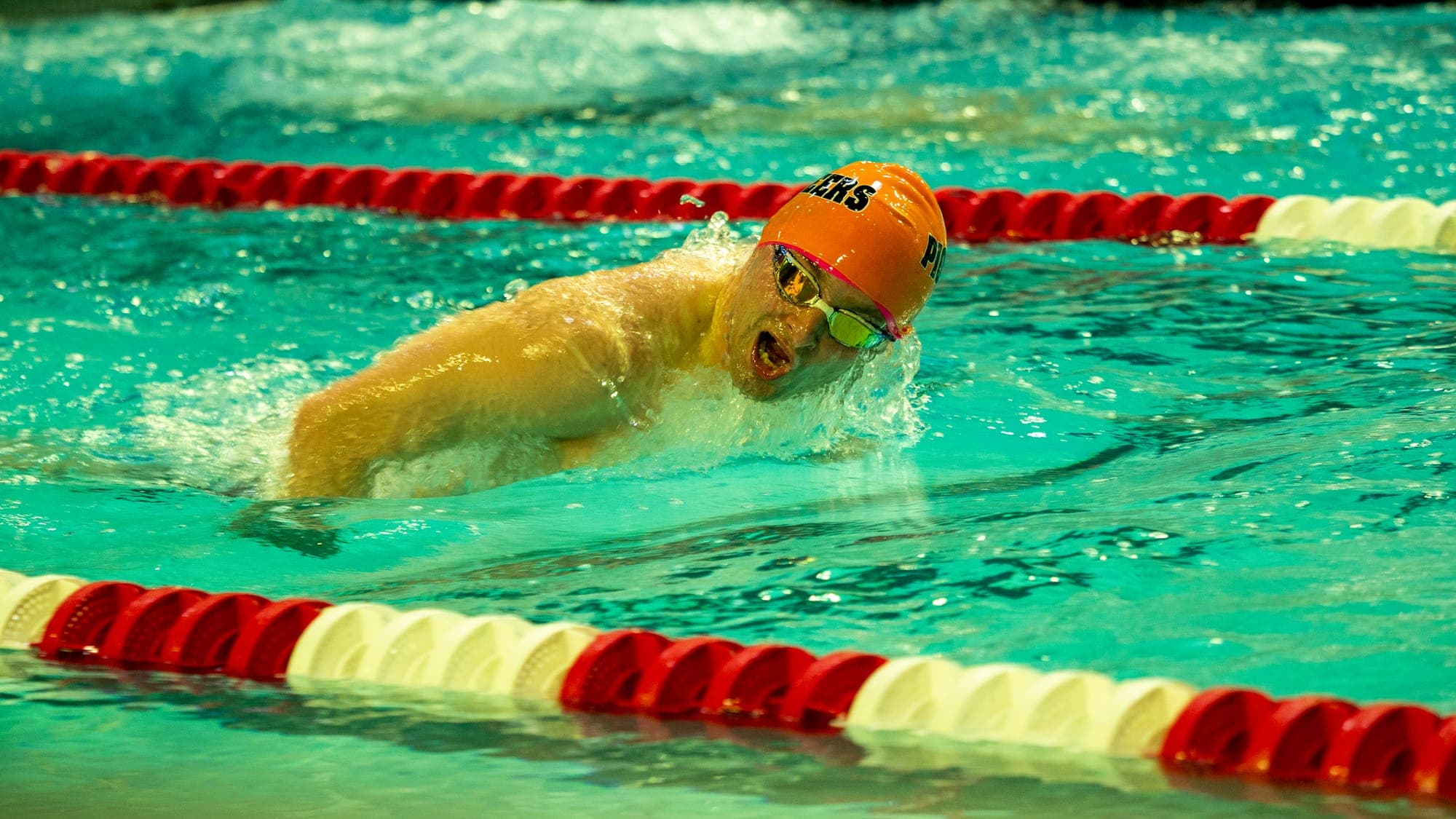 Swimmer in an orange cap mid-butterfly stroke, mouth open for air as water sheets off his shoulders