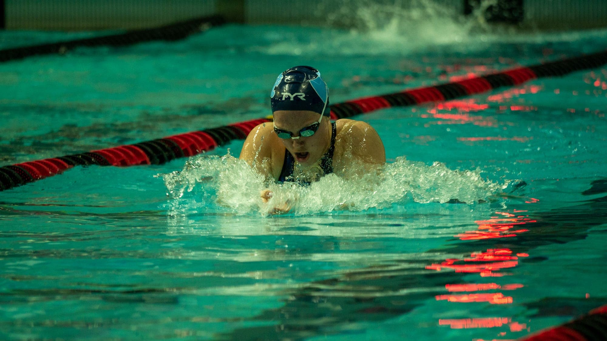 Swimmer in a TYR cap surging forward in breaststroke, red lights reflecting on the churned green water
