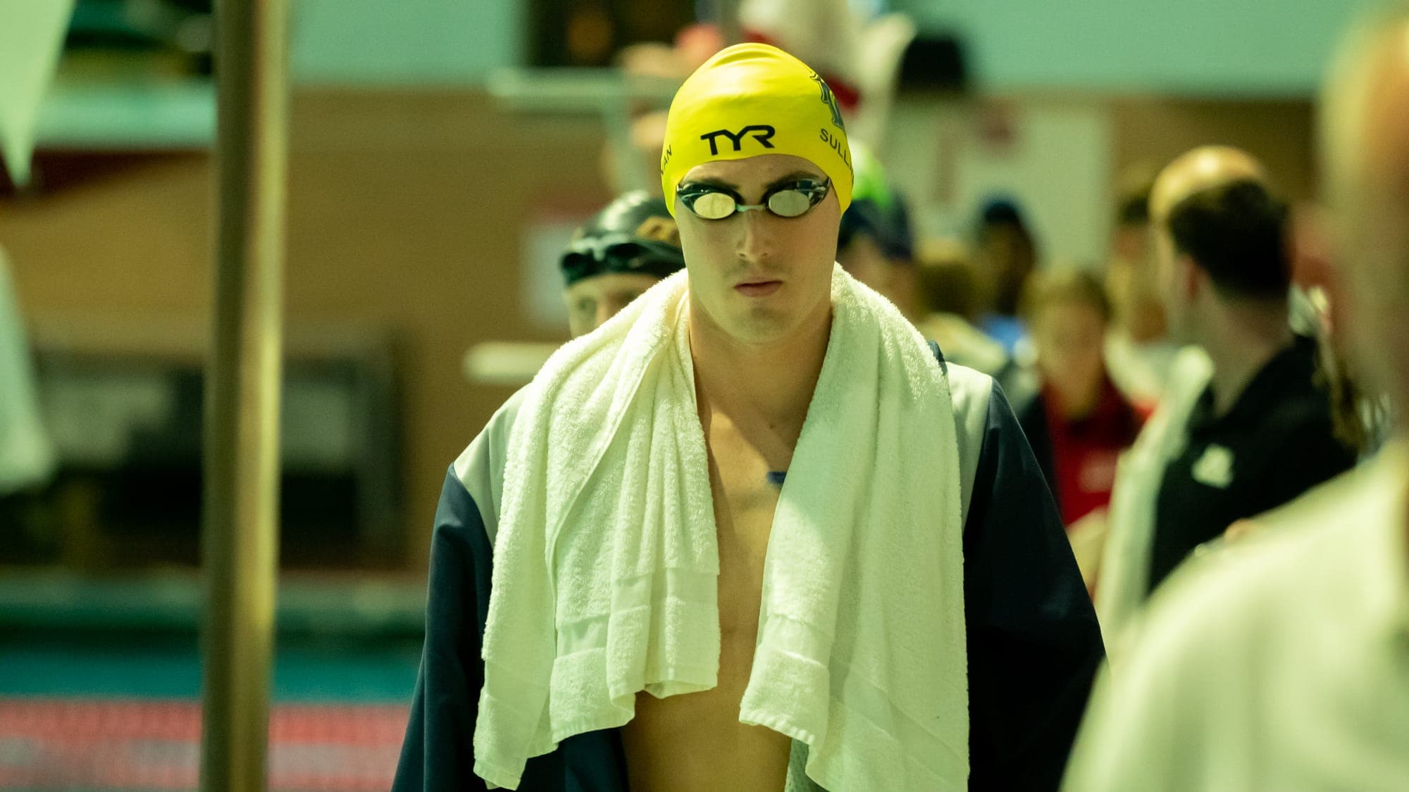 Swimmer draped in a white towel, yellow cap and mirrored goggles catching the light as he walks off the deck
