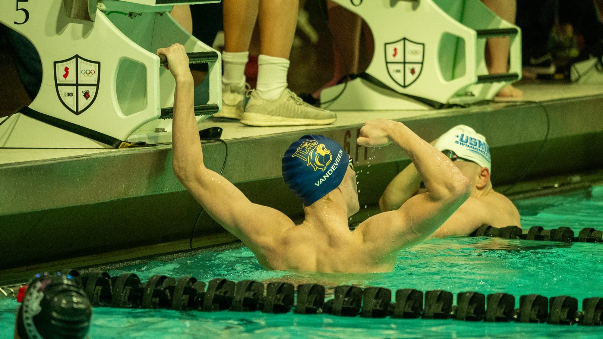 Swimmer at the pool wall with both arms raised in celebration beneath the starting blocks, a rival looking on
