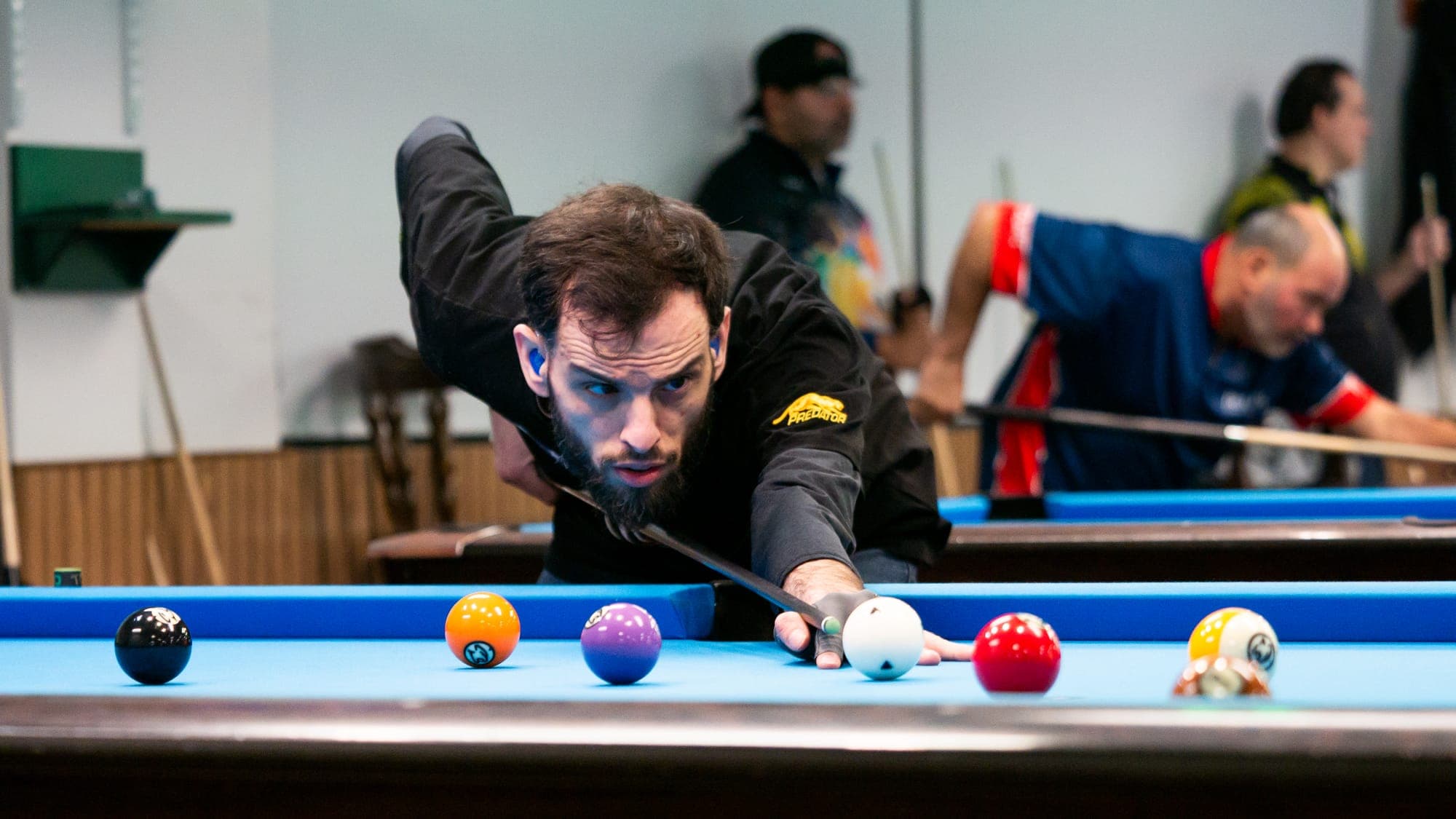 Billiards player sighting down his cue at the white ball on a blue-felt table, rivals bent over tables behind him