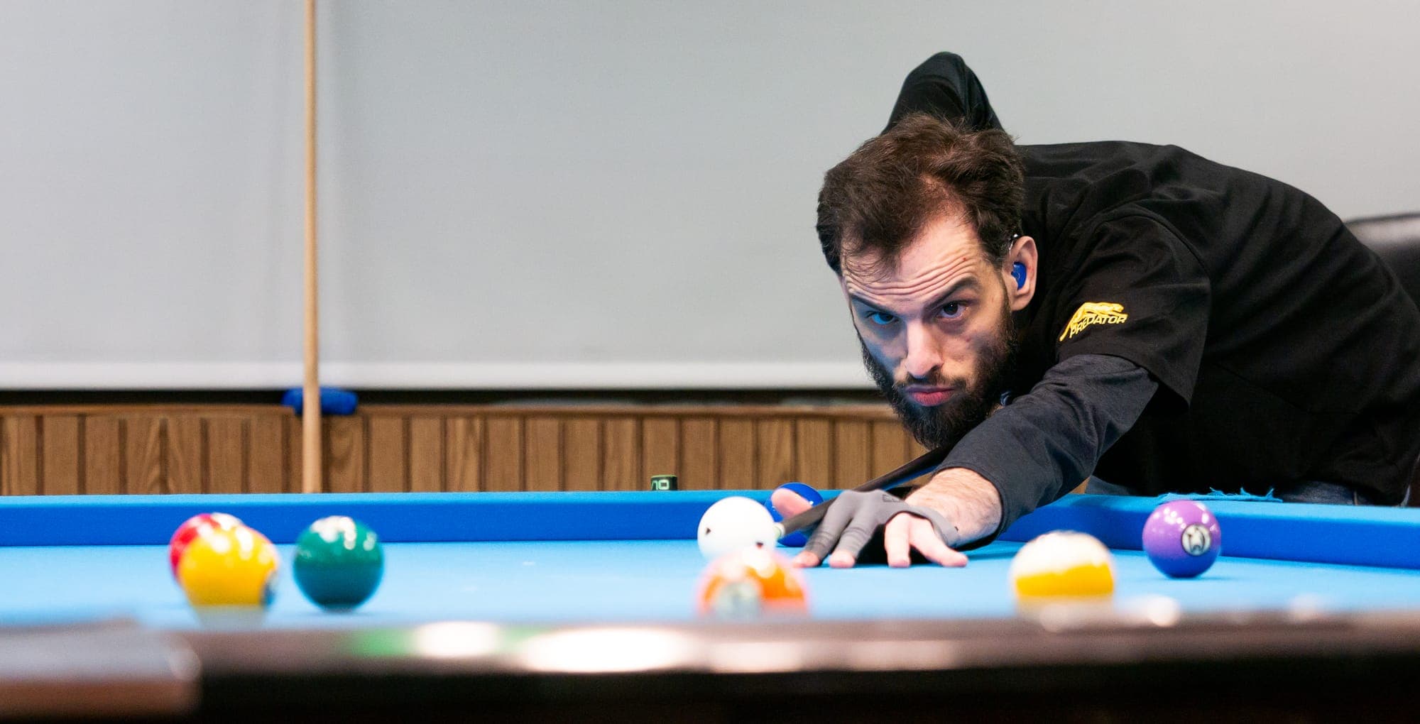 Bearded pool player in black stretched across the table, eyes fixed past the cue ball amid scattered colored balls