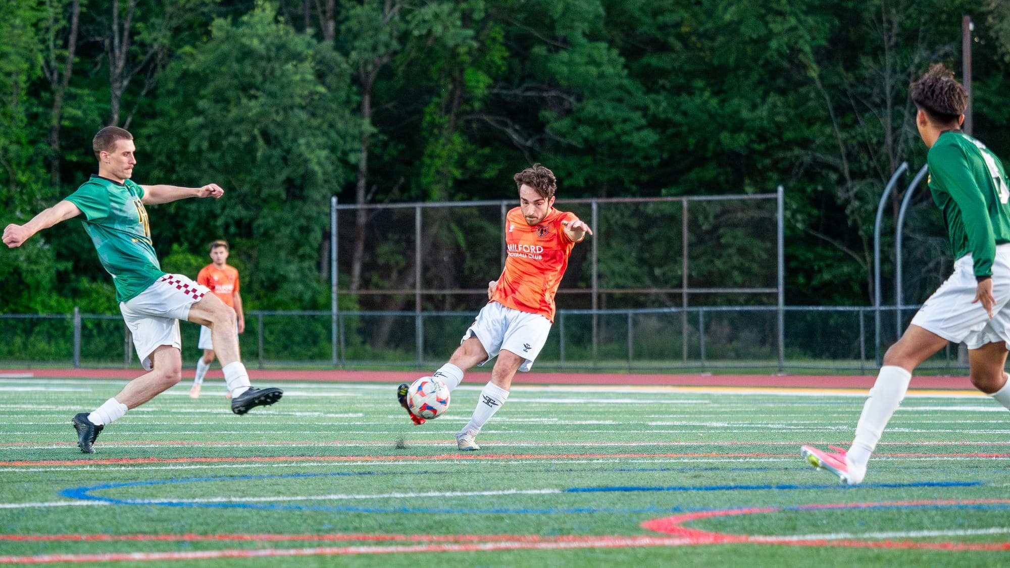 Soccer player in orange firing a shot as two defenders in green stretch to close him down
