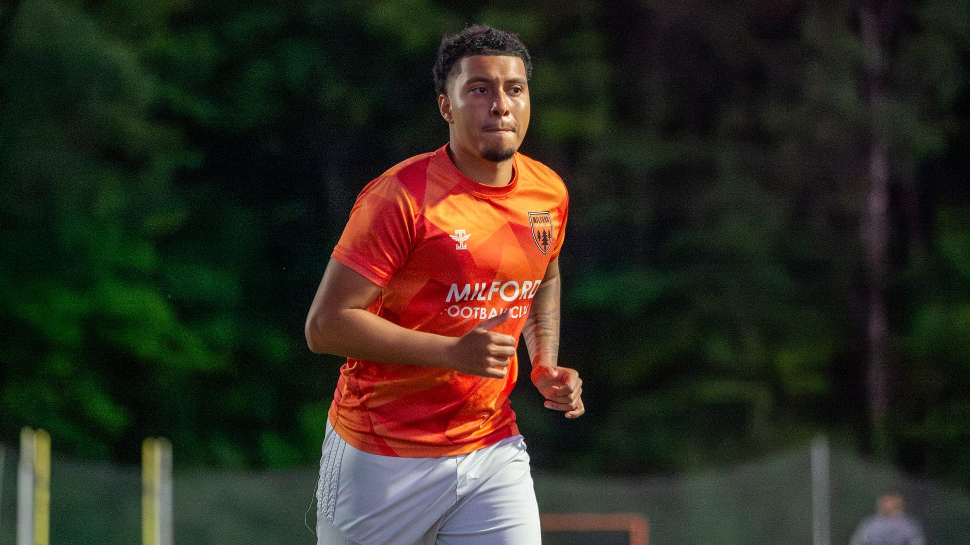Close view of a Milford FC player jogging, eyes fixed downfield against dark trees