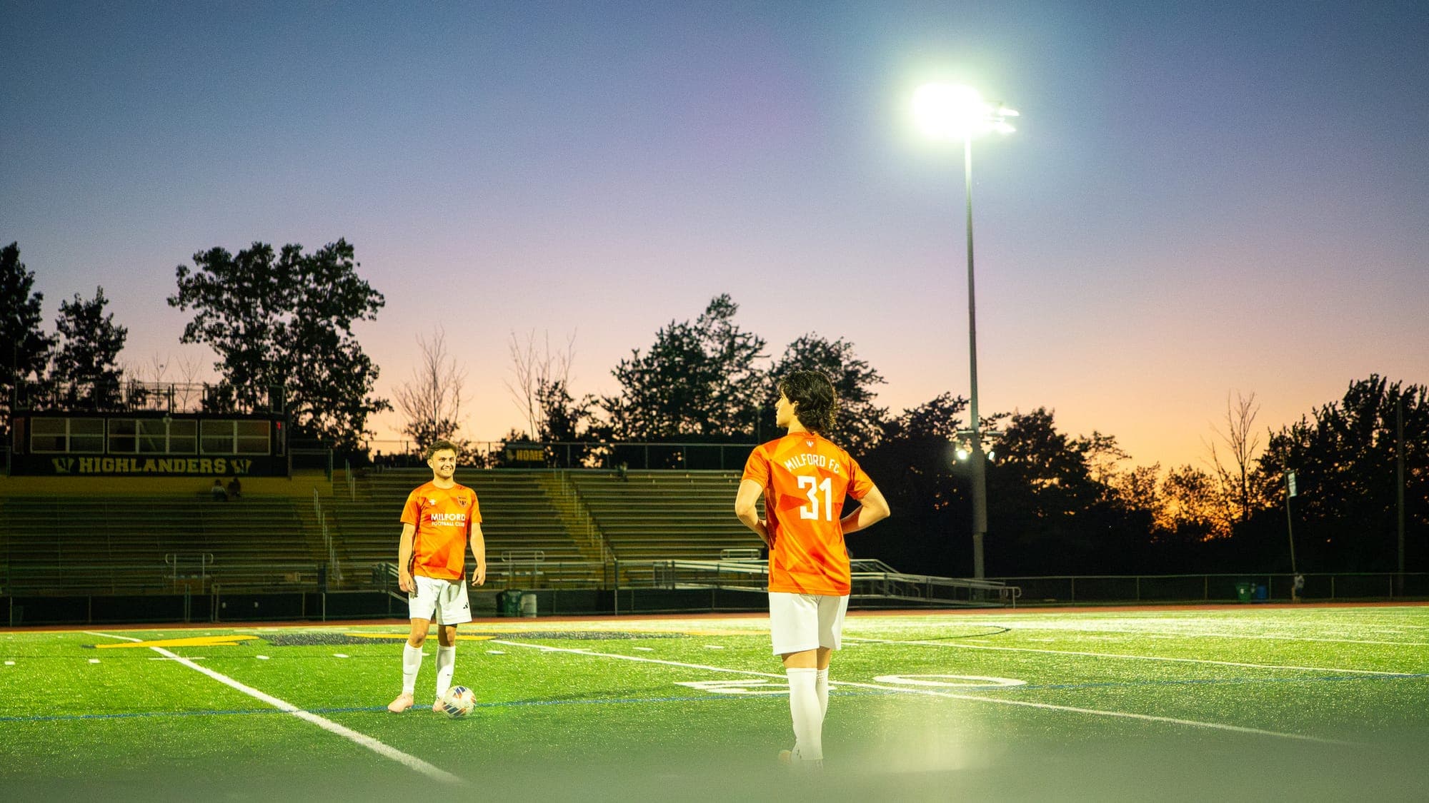 Two soccer players waiting on a floodlit field at dusk, empty bleachers under a fading orange sky
