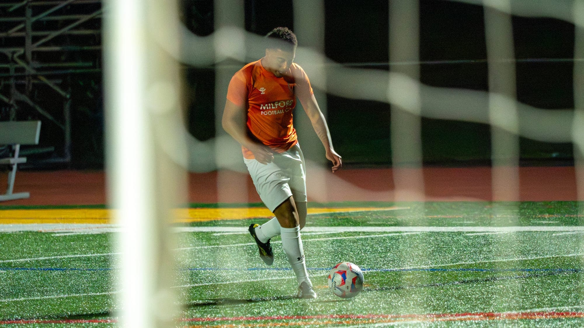 Soccer player lining up a shot on glistening night turf, seen through the goal netting